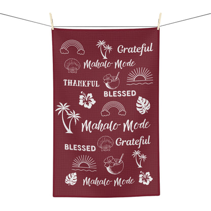 A rectangular waffle-weave kitchen towel in deep burgundy with white tropical-themed graphics and phrases like 'Grateful', 'Blessed', and 'Mahalo Mode', hanging from a clothesline with wooden clips against a white background.
