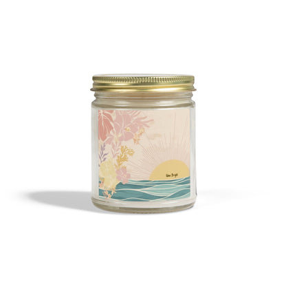 A glass jar candle with a gold metal screw-on lid and a pastel illustrated label depicting a sunset over waves with tropical flowers. The label has the text 'sea-breeze' on it.