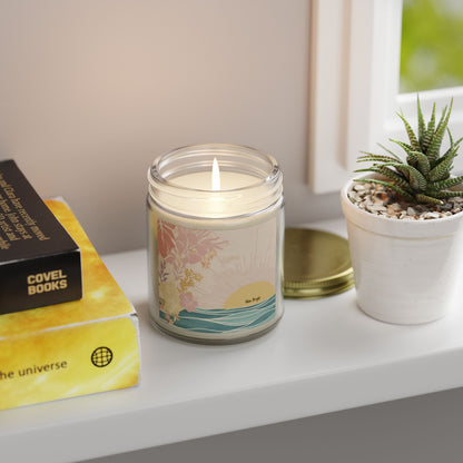 A lit glass jar candle with a colorful tropical sunset label sits on a white windowsill next to a small potted succulent and a gold lid. A yellow and black book is partially visible on the left.