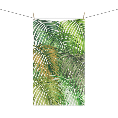 A rectangular tea towel featuring a watercolor tropical palm leaf pattern in shades of green and yellow, hanging from a string with two wooden clothespins against a plain white background.