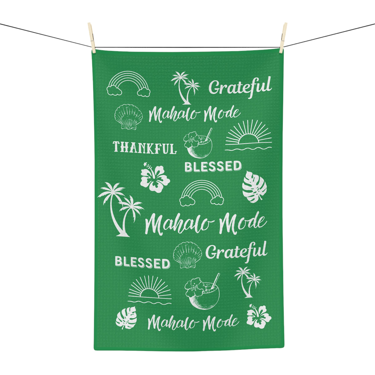 A green kitchen towel or tea towel hanging from a clothesline with two wooden clips, featuring white tropical-themed illustrations and phrases including 'Mahalo Mode,' 'Grateful,' 'Blessed,' 'Thankful,' palm trees, hibiscus flowers, monstera leaves, rainbows, shells, and sunsets.