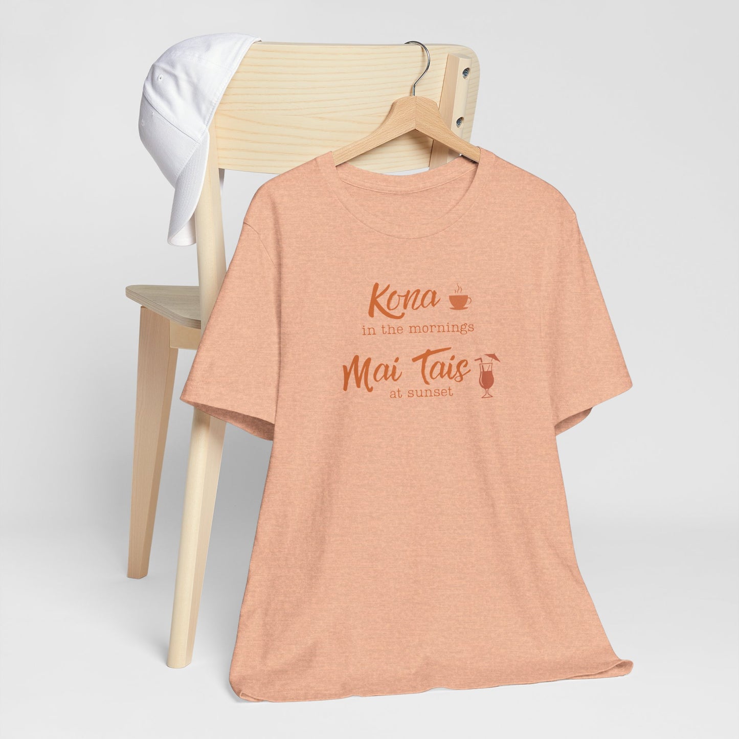 A peach heather graphic t-shirt hanging on a wooden hanger, styled in front of a light wood chair with a white towel draped over the backrest. The shirt features a coral orange graphic that reads 'Kona in the mornings Mai Tais at sunset' with small coffee and cocktail illustrations.