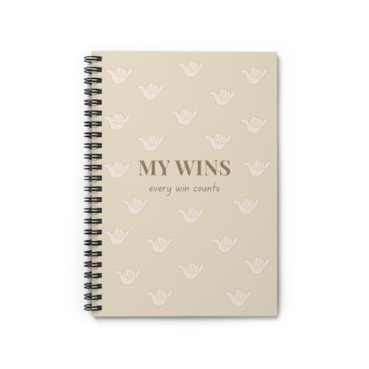 A beige spiral-bound notebook with a black wire binding, featuring the text 'MY WINS every win counts' and a repeating pattern of white shaka icons on the cover, shown against a plain white background.