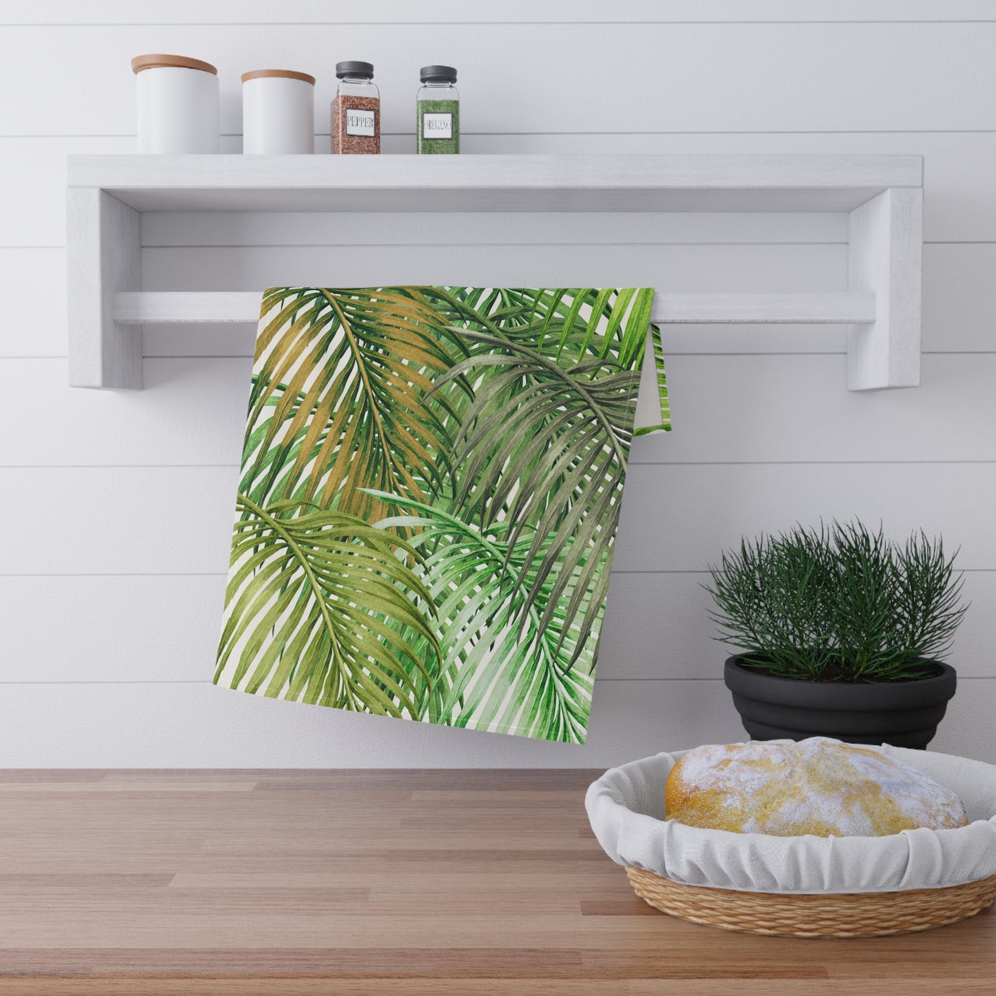 A kitchen towel with a green palm leaf pattern hanging from a white wall-mounted rack in a bright kitchen setting.