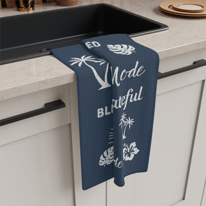 A navy blue kitchen towel with white tropical and text graphics hangs over the edge of a black sink in a modern kitchen setting.