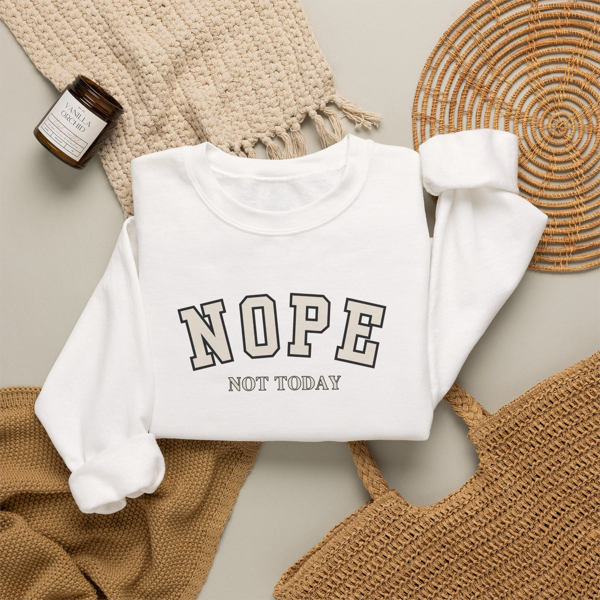 A folded white crewneck sweatshirt with the text 'NOPE NOT TODAY' printed on the front, styled in a flat lay with a beige knit blanket, a woven basket, a small candle, and a tan tote bag on a light gray background.