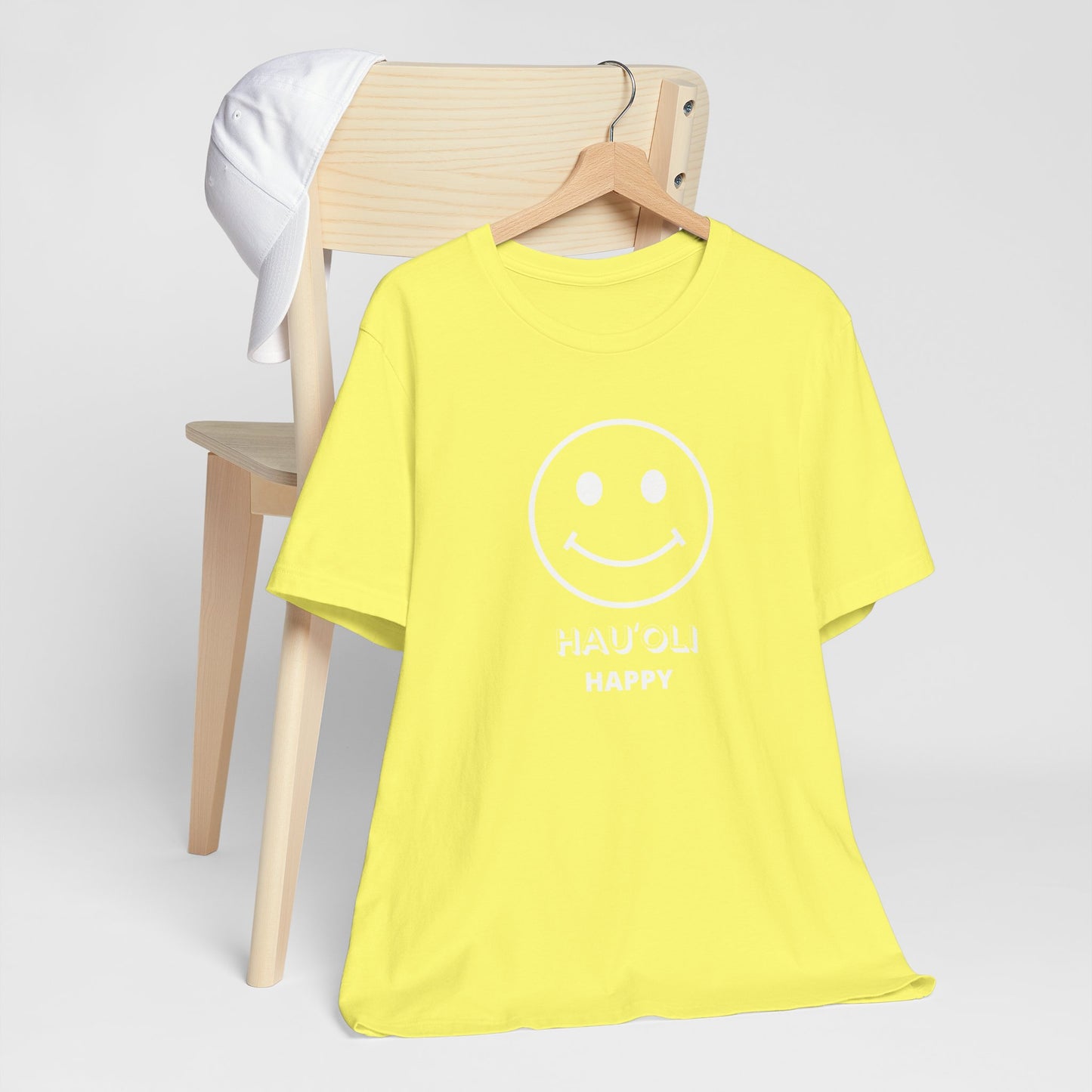 A bright yellow short-sleeve t-shirt with a white smiley face graphic and the text 'HAUʻOLI HAPPY' hanging on a wooden hanger, displayed in front of a light wood chair with a white shirt draped over it, against a light gray background.