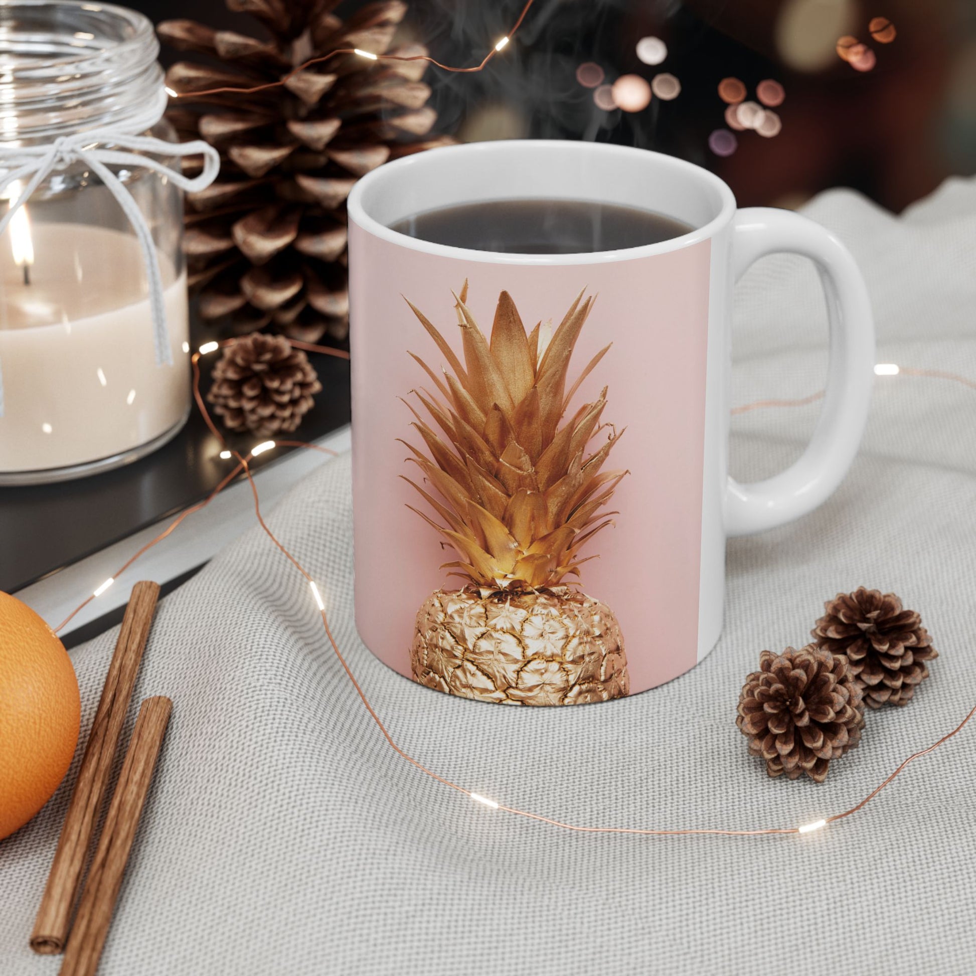 A white ceramic mug with a light pink front panel featuring a metallic gold pineapple design, filled with black coffee and styled on a light fabric surface with pinecones, fairy lights, cinnamon sticks, and a candle.