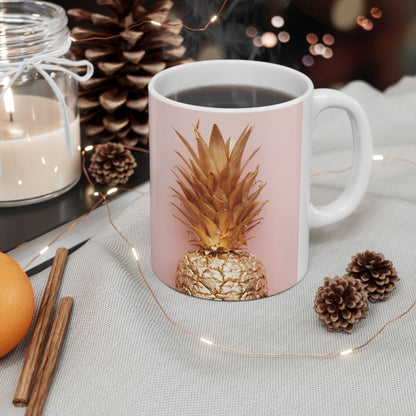 A white ceramic mug with a light pink front panel featuring a metallic gold pineapple design, filled with black coffee and styled on a light fabric surface with pinecones, fairy lights, cinnamon sticks, and a candle.