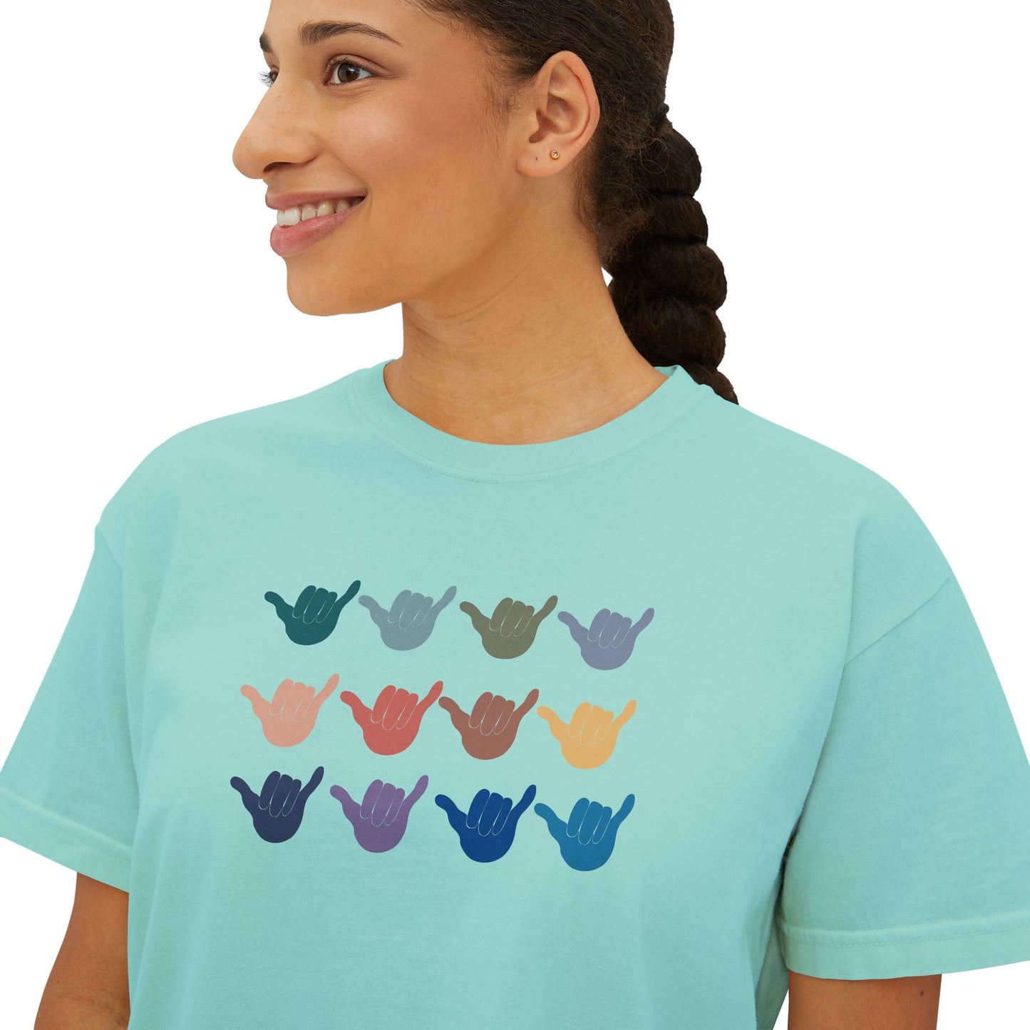 A woman wearing a light aqua blue t-shirt with a graphic of twelve shaka signs in various colors arranged in a grid. The background is solid white.