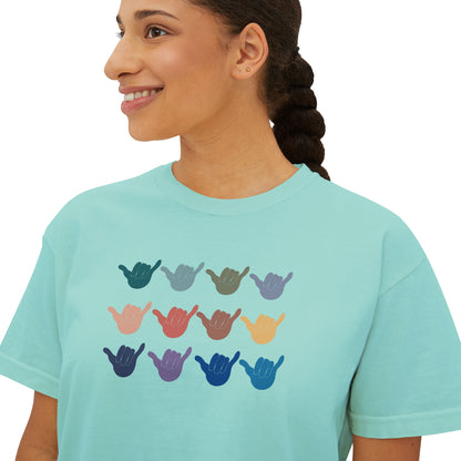 A woman wearing a light aqua blue t-shirt with a graphic of twelve shaka signs in various colors arranged in a grid. The background is solid white.