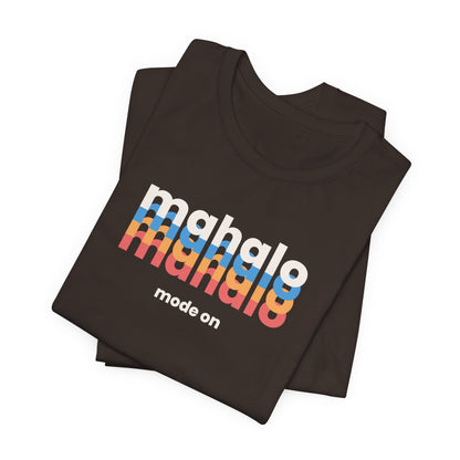 A folded dark brown t-shirt with a retro-style graphic reading 'mahalo mahalo' in white, blue, orange, and red gradient letters, with 'mode on' printed below in white, on a clean white background.