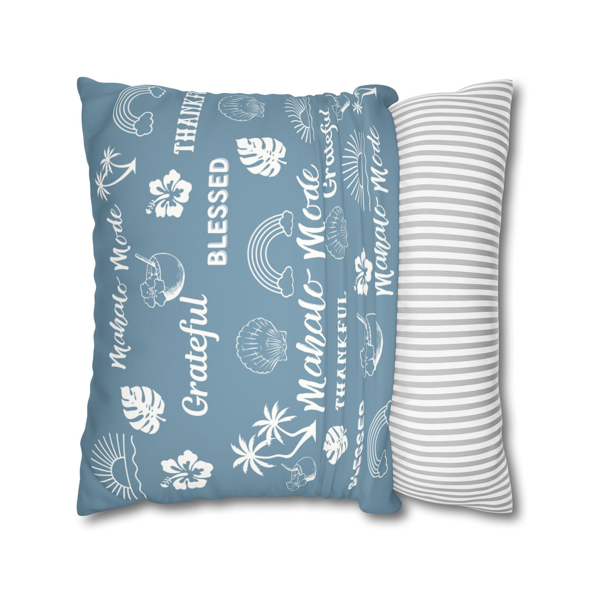 A rectangular throw pillow in dusty blue with white tropical beach-themed illustrations and text including 'Mahalo - Mo'lelo', 'Blessed', and 'Grateful', shown next to a white pillow with thin gray stripes on a white background.