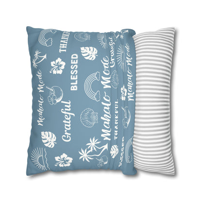 A rectangular throw pillow in dusty blue with white tropical beach-themed illustrations and text including 'Mahalo - Mo'lelo', 'Blessed', and 'Grateful', shown next to a white pillow with thin gray stripes on a white background.