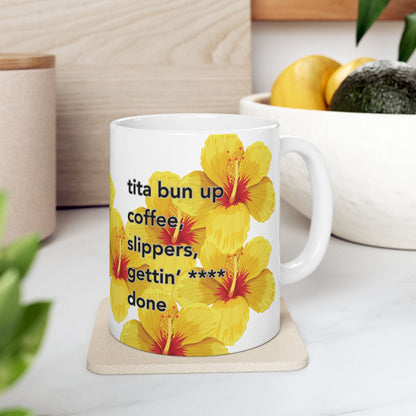 Tita Bun coffee mug with yellow hibiscus design by Global Village Kailua sitting on a wood coaster on a white kitchen with a white bowl of fruit behind it.