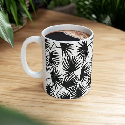 A white ceramic mug with a black palm leaf pattern, filled with black coffee and set on a light wood table with green plants in the background.