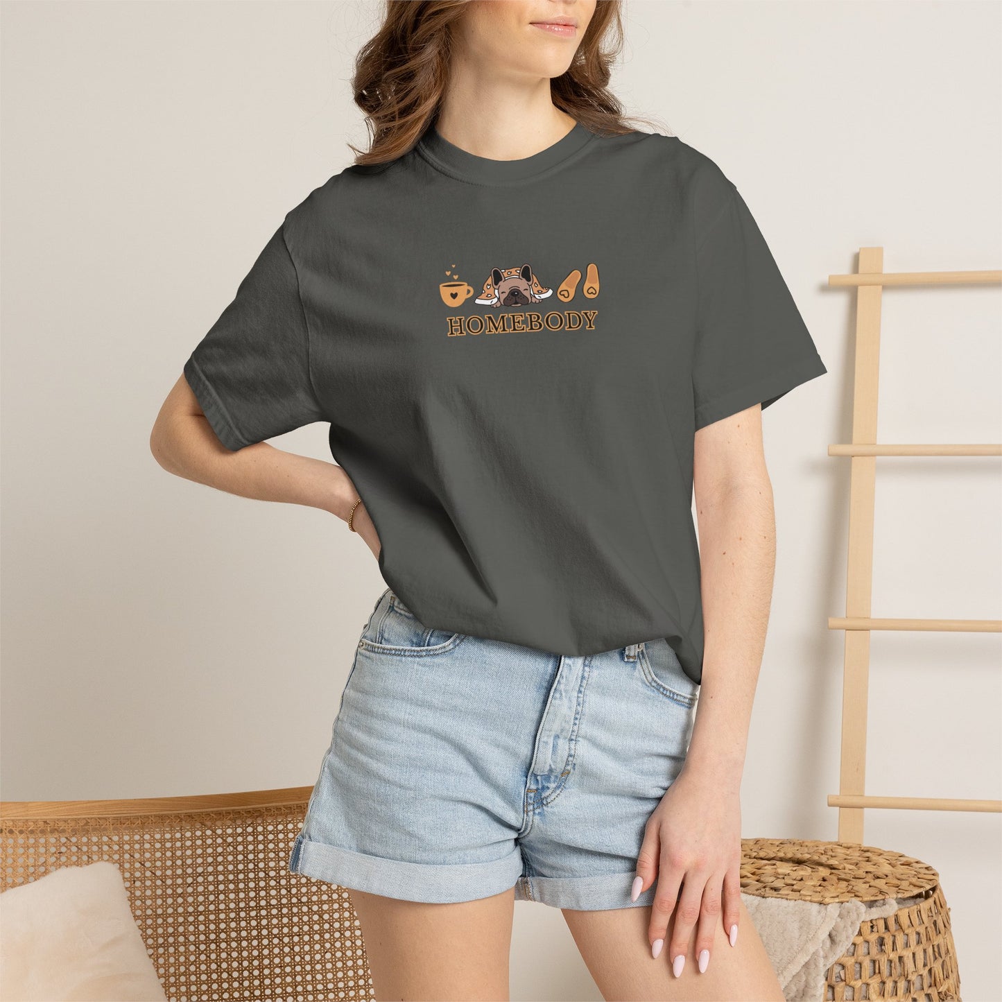 Person wearing a dark short-sleeve crewneck t-shirt with a graphic on the chest depicting a dog under a blanket, a coffee mug, a pair of house slippers, and the word 'HOMEBODY' in brown text. Standing wearing denim shorts.