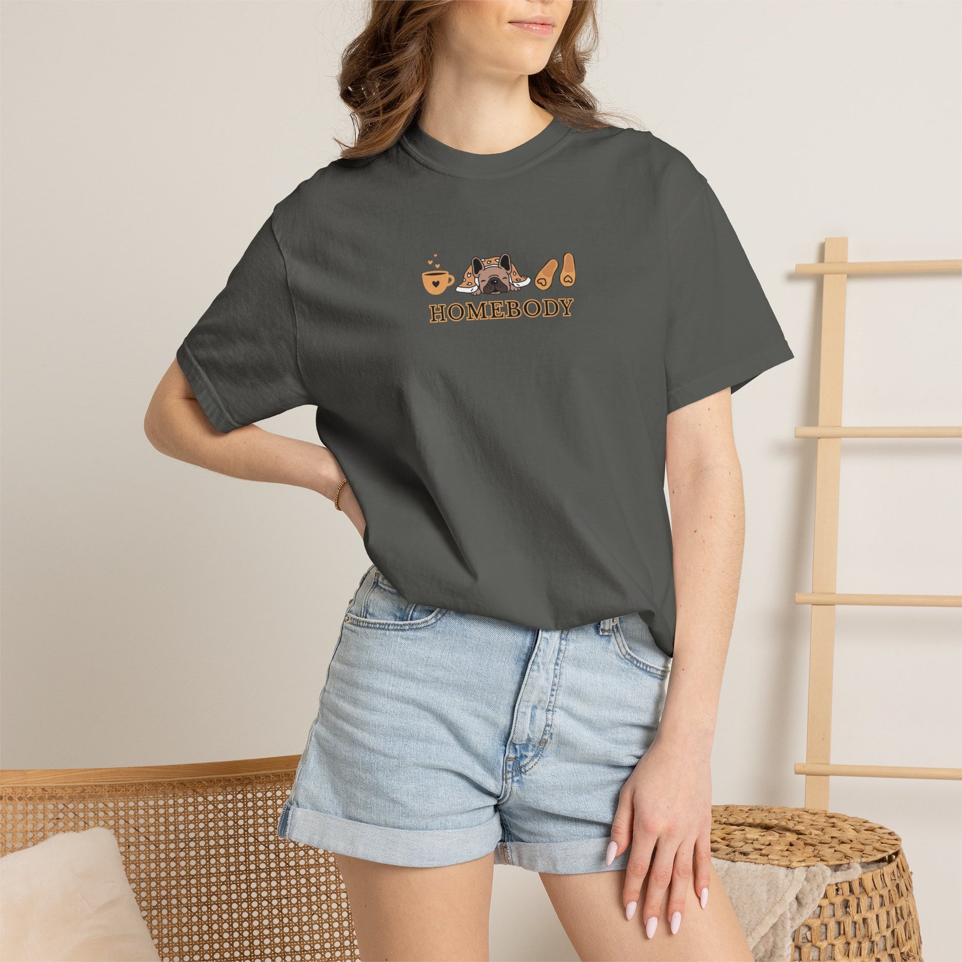 Person wearing a dark short-sleeve crewneck t-shirt with a graphic on the chest depicting a dog under a blanket, a coffee mug, a pair of house slippers, and the word 'HOMEBODY' in brown text. Standing wearing denim shorts.