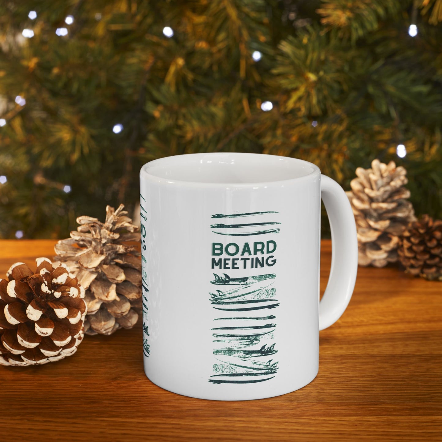 A white ceramic mug with a green 'BOARD MEETING' graphic sits on a wooden table surrounded by pinecones, with a blurred Christmas tree in the background.