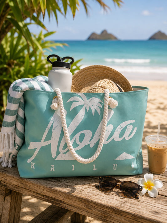A turquoise tote bag with white rope handles and a white palm tree graphic reading 'Aloha Kailua' sits on a beach picnic table. A straw hat, white towel, water bottle, sunglasses, iced coffee, and flower are arranged around it, with ocean and palm trees in the background.