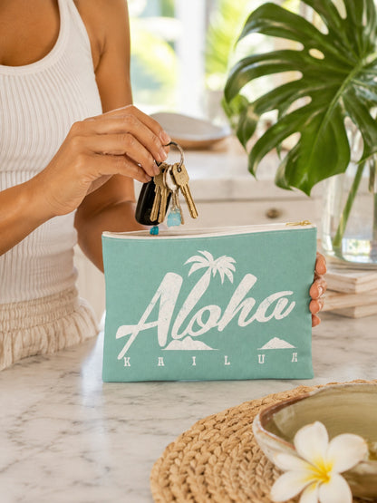 A person in a white ribbed tank top holds a mint green zip pouch with a white 'Aloha Kailua' palm tree graphic, inserting keys into the pouch on a marble countertop with tropical plant decor in the background.
