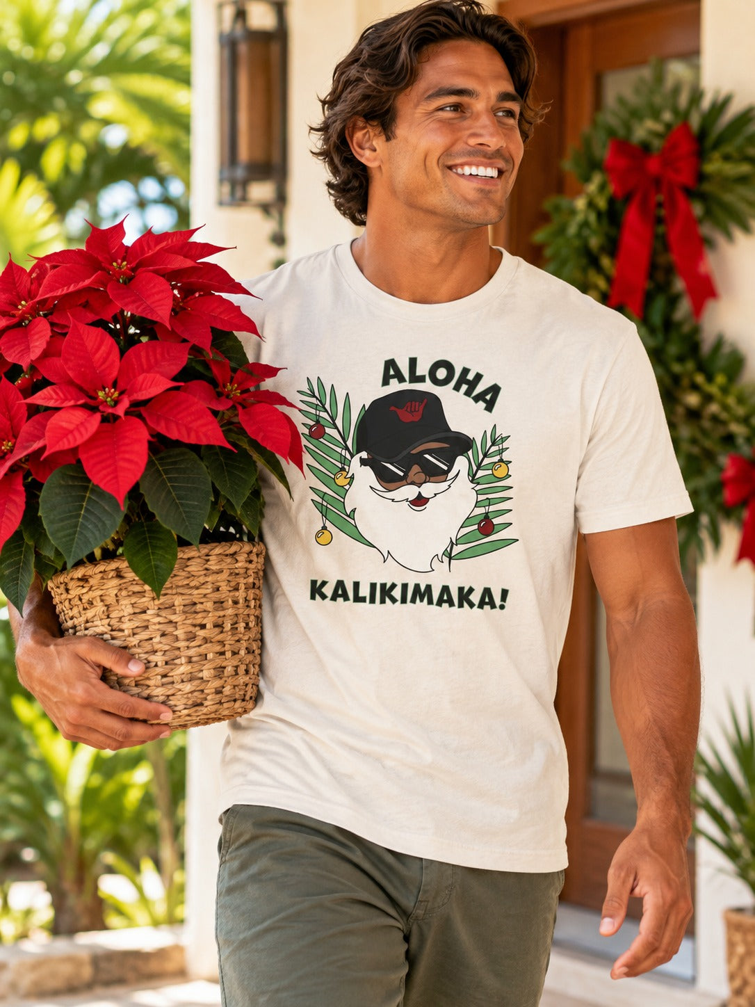 Man wearing white Aloha Kalikimaka Santa graphic tee carrying a red poinsettia on a tropical holiday porch.