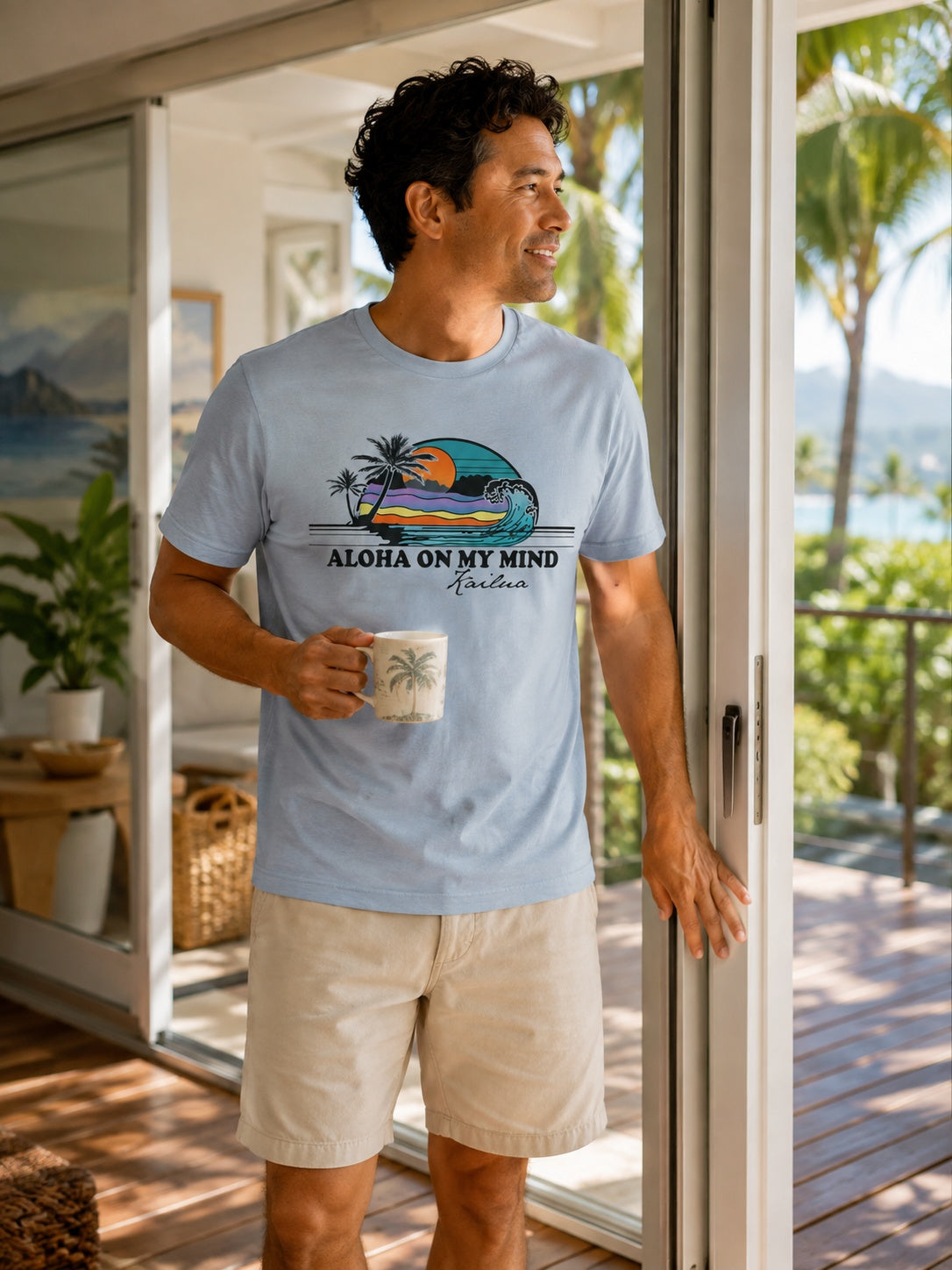 A man wearing a light blue graphic t-shirt with 'ALOHA ON MY MIND Hawaii Kailua' printed on the front, standing on a wooden deck in front of sliding glass doors with a tropical ocean view.