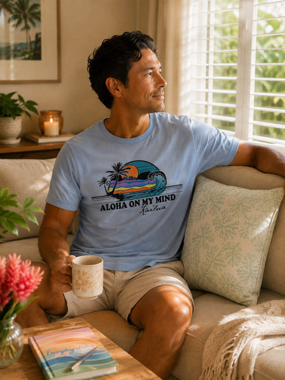 A man sitting on a beige couch in a sunlit living room, wearing a light blue graphic T-shirt with a tropical beach scene and the text 'ALOHA ON MY MIND' and 'Kailua'.