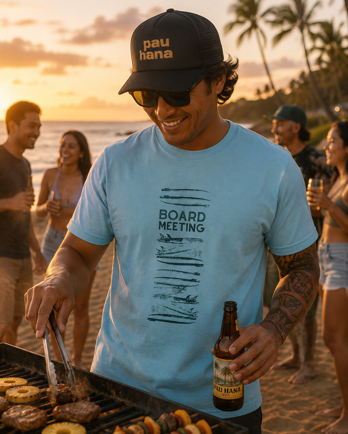 Man grilling food on a beach with sunset and ocean view, wearing a 'pau hana' cap and 'Board Meeting' t-shirt.