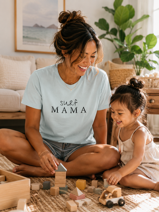 Mom wearing the Surf Mama graphic tee while playing with her young daughter in a bright, coastal-inspired home.
