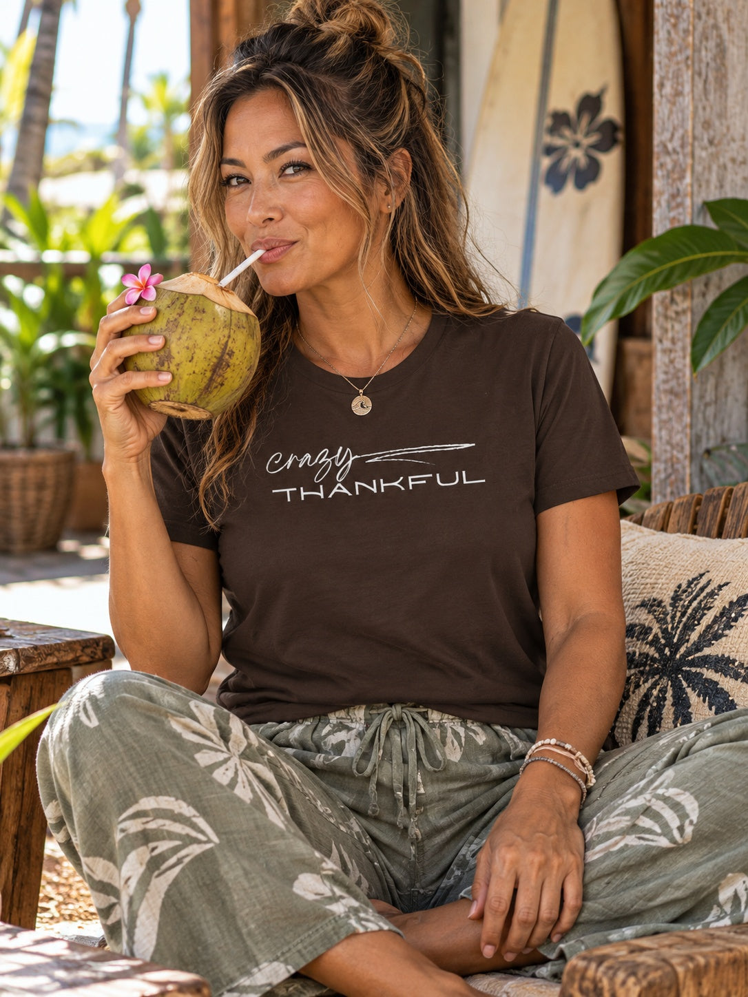 A woman sitting outdoors on a porch wearing a dark brown graphic t-shirt with 'crazy THANKFUL' printed in white, paired with green and white palm-patterned pants. She is holding a coconut drink with a flower garnish.
