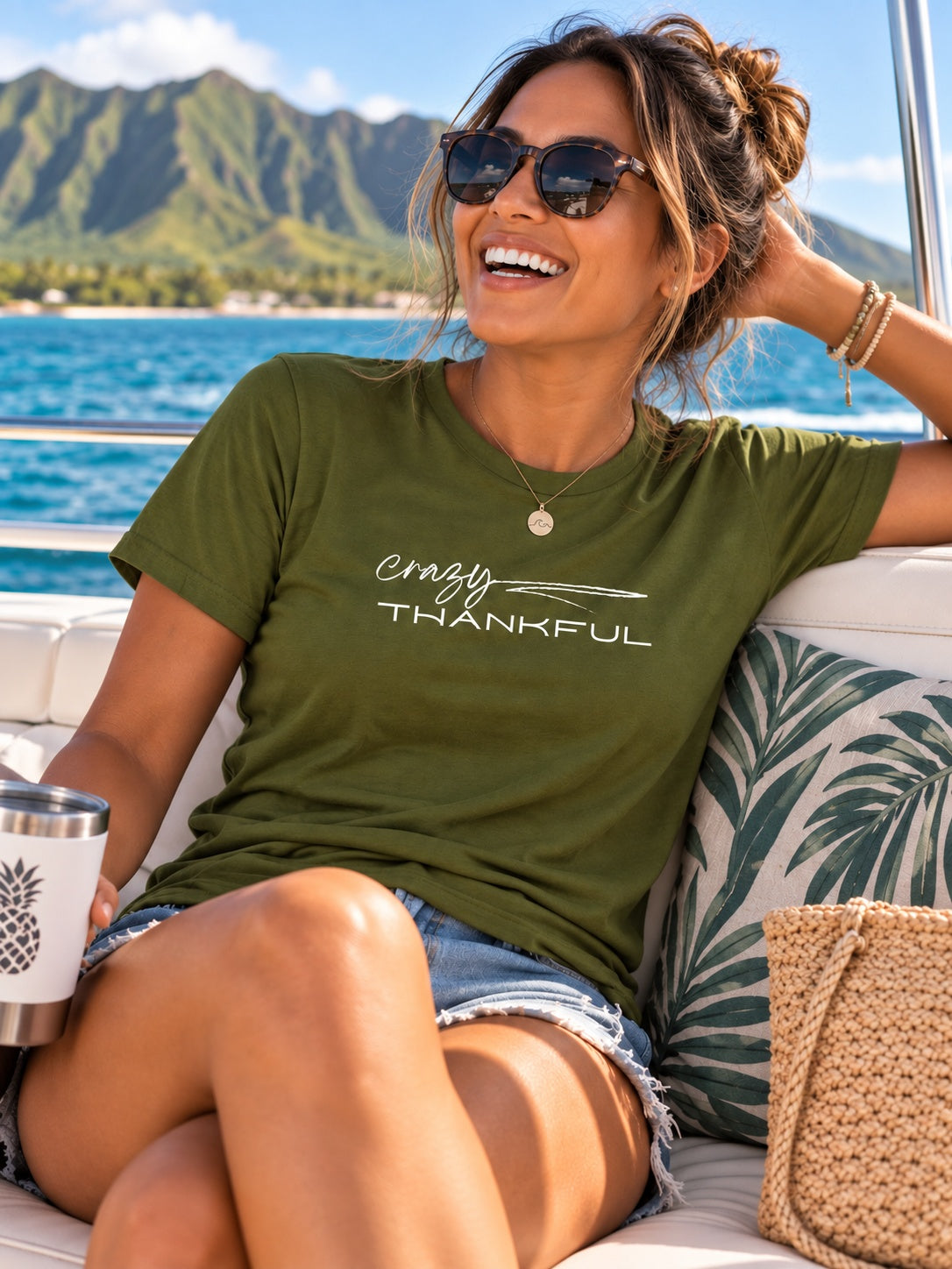 A woman wearing an olive green graphic t-shirt with the words 'Crazy Thankful' in white script and block lettering, sitting on a boat with a tropical ocean and mountain backdrop.