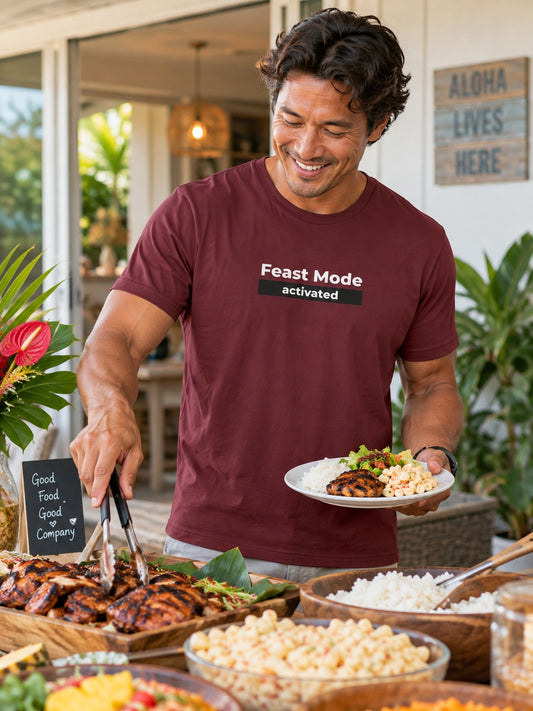 A smiling man in a maroon t-shirt with 'Feast Mode activated' printed on the chest is serving food from a buffet table in a tropical outdoor setting.