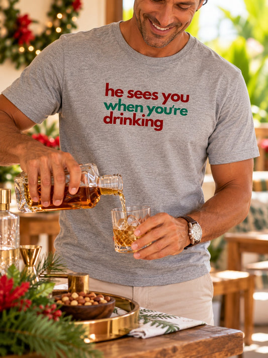 Man wearing a gray He Sees You When You're Drinking Christmas tee pouring whiskey at a holiday gathering.