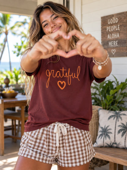 A woman posing on a tropical porch wearing a maroon t-shirt with 'grateful' and a heart printed in orange, paired with brown and white gingham shorts.