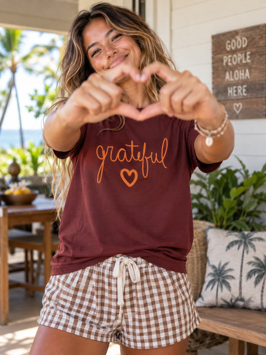 A woman posing on a tropical porch wearing a maroon t-shirt with 'grateful' and a heart printed in orange, paired with brown and white gingham shorts.