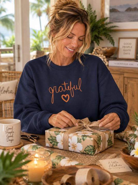 A woman in a navy blue sweatshirt with the word 'grateful' and a small heart in orange on the front, wrapping a floral gift box with a twine bow in a warm, decorated home setting.