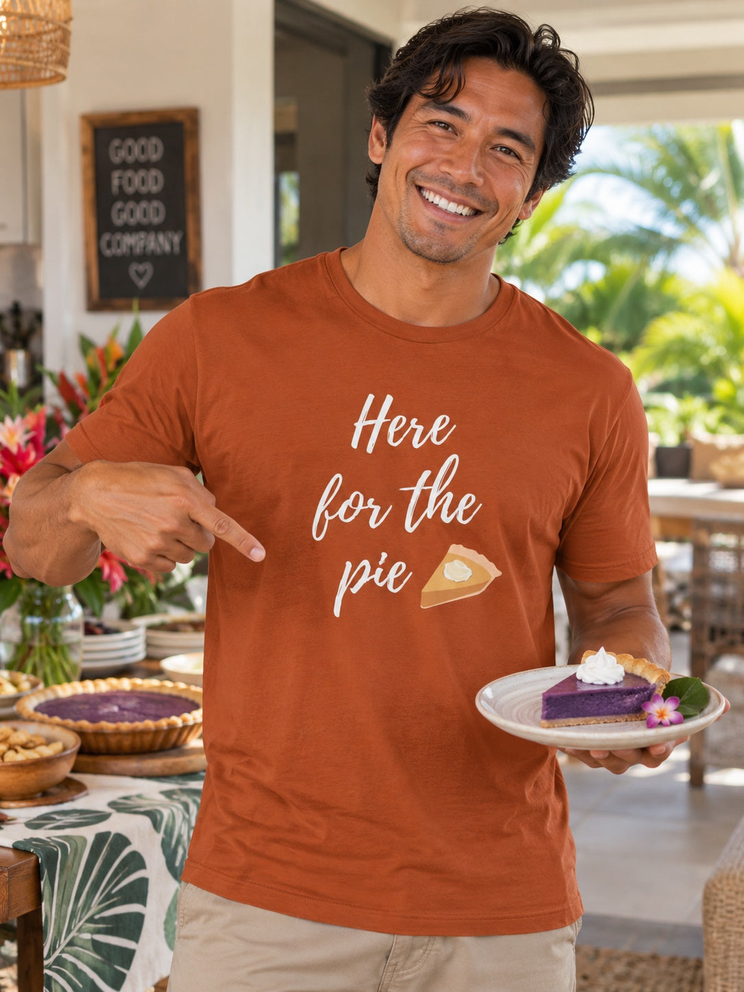 A smiling man wearing a rust-orange graphic t-shirt that reads 'Here for the pie' with a slice of pie illustration, holding a plate of purple pie in a tropical outdoor patio setting.