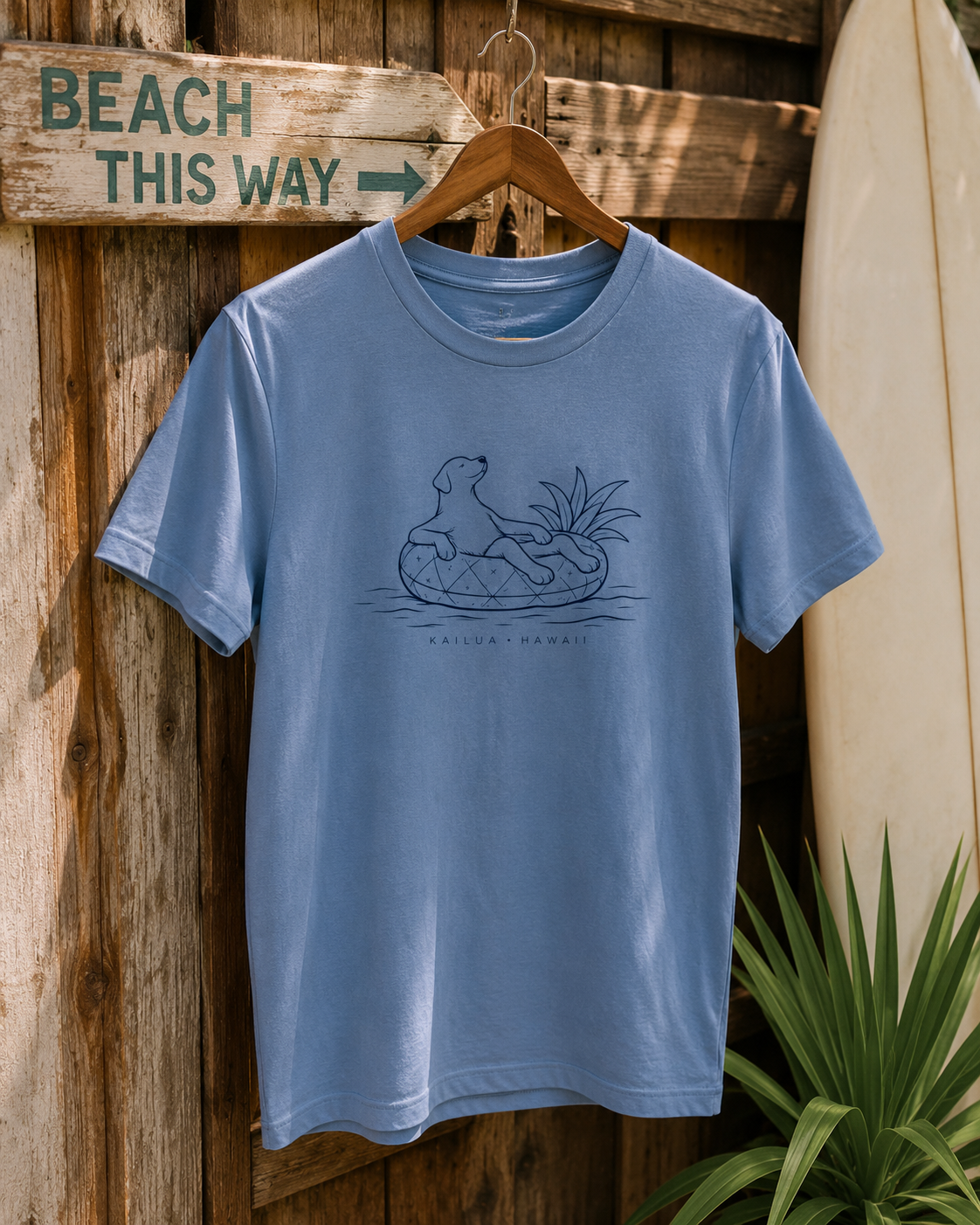 A light blue crewneck t-shirt hanging on a wooden hanger against a rustic wooden wall. The shirt features a dark blue line art graphic of a dog sitting in a pineapple floatie, with the text 'KAILUA HAWAII' printed below.