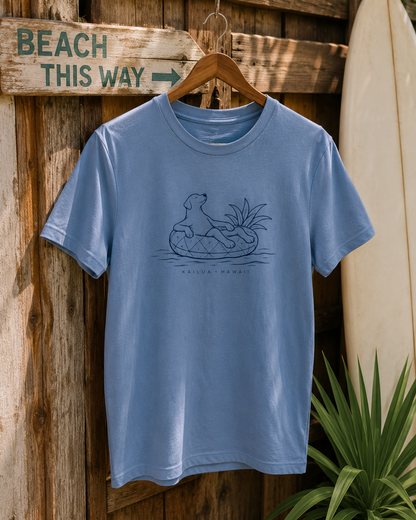 A light blue crewneck t-shirt hanging on a wooden hanger against a rustic wooden wall. The shirt features a dark blue line art graphic of a dog sitting in a pineapple floatie, with the text 'KAILUA HAWAII' printed below.