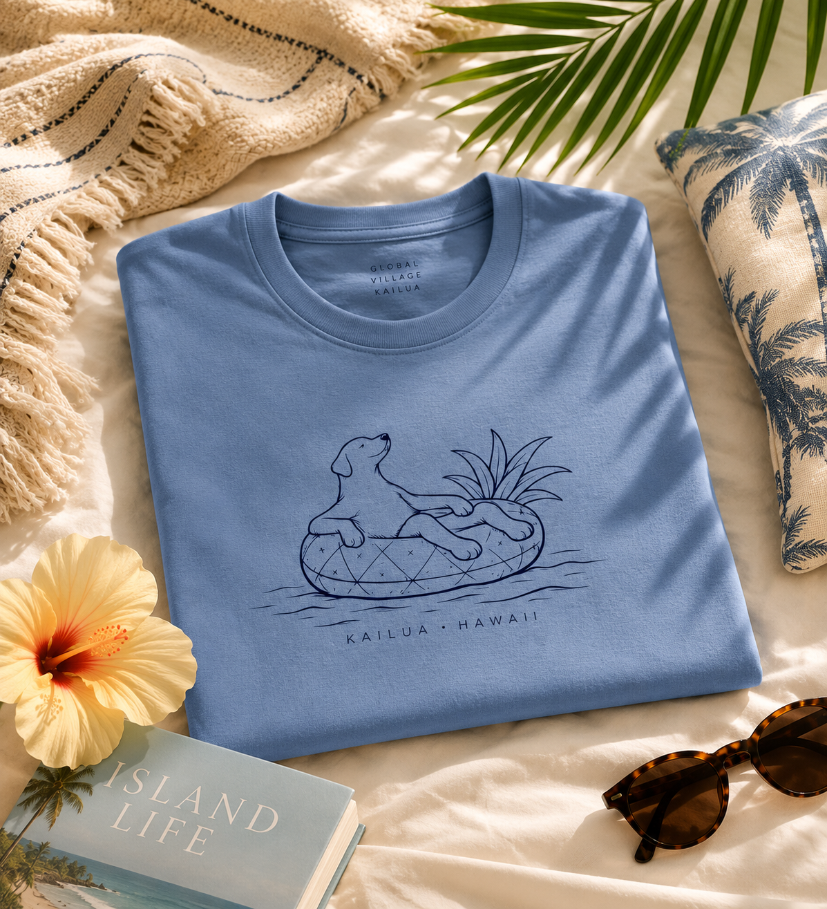 A folded light blue T-shirt with a navy line-art graphic of a dog lounging on a pineapple float, with the text 'KAILUA - HAWAII' below. The shirt is styled in a beach-themed flat lay on a cream fabric surface with a palm leaf, sunglasses, a hibiscus flower, a book, and a patterned pillow.