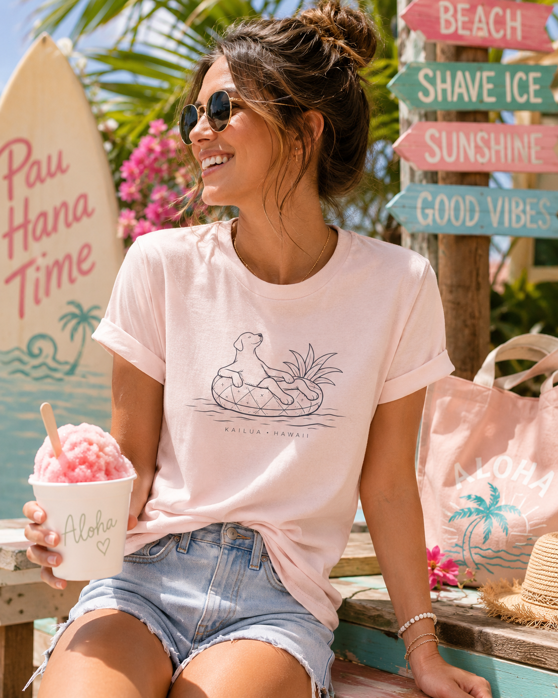 A woman wearing a light pink graphic t-shirt with a dog on pineapple floatie design and 'Kailua Hawaii' text, sitting outdoors on a beach-themed bench with a drink and sunglasses.