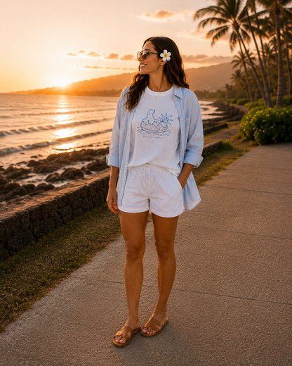 A woman stands on a seaside walkway at sunset wearing a white graphic T-shirt ʻKoa Dog on Pineapple Floatieʻ white shorts, an open light blue button-up shirt, and tan sandals. Palm trees and ocean waves are visible in the background.