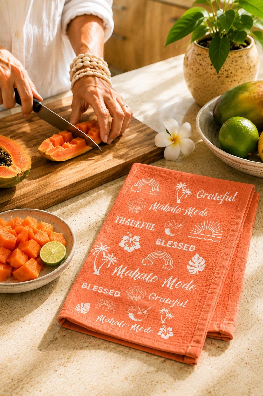 An orange kitchen towel with white tropical and Hawaiian-themed text and illustrations, laid on a countertop next to a person slicing papaya. The towel features phrases like 'Grateful', 'Mahalo Mode', and 'Blessed'.