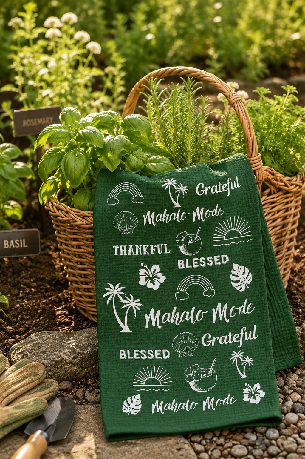 A dark green waffle-weave kitchen towel with white tropical and beach-themed text and graphic prints, styled in a garden setting next to a wicker basket of fresh herbs.
