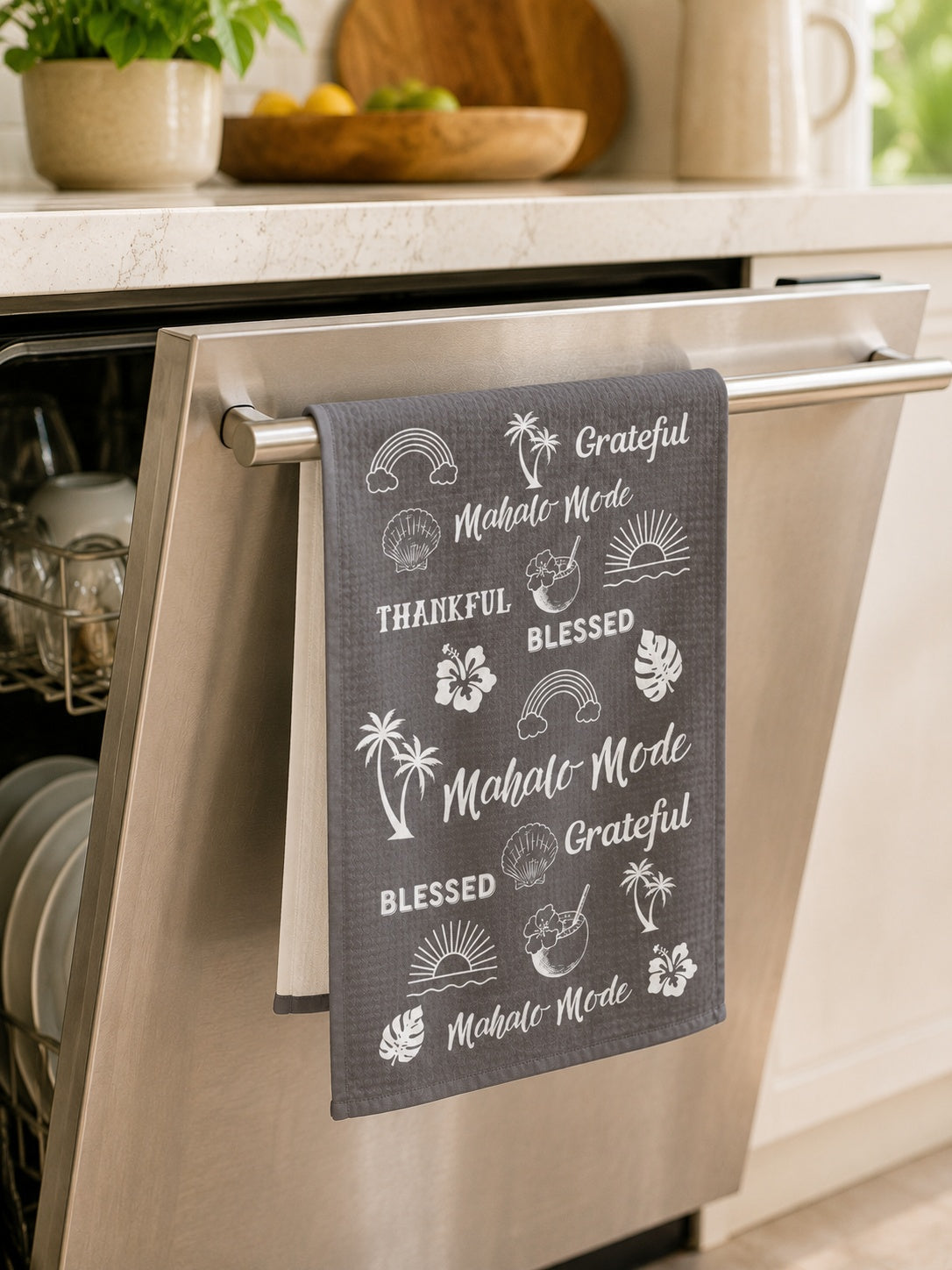 A dark gray kitchen towel with white tropical and beach-themed illustrations and phrases like 'Grateful', 'Mahalo Mode', and 'Blessed' hanging from the handle of a stainless steel dishwasher in a bright kitchen.