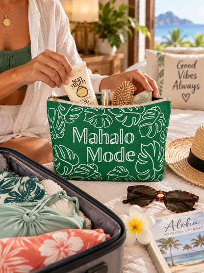 A green toiletry bag with a white tropical leaf pattern and the words 'Mahalo Mode' printed on the front, being opened by a person in a beach-themed setting. The scene includes a suitcase with tropical towels, sunglasses, a straw hat, a flower, and a travel book.