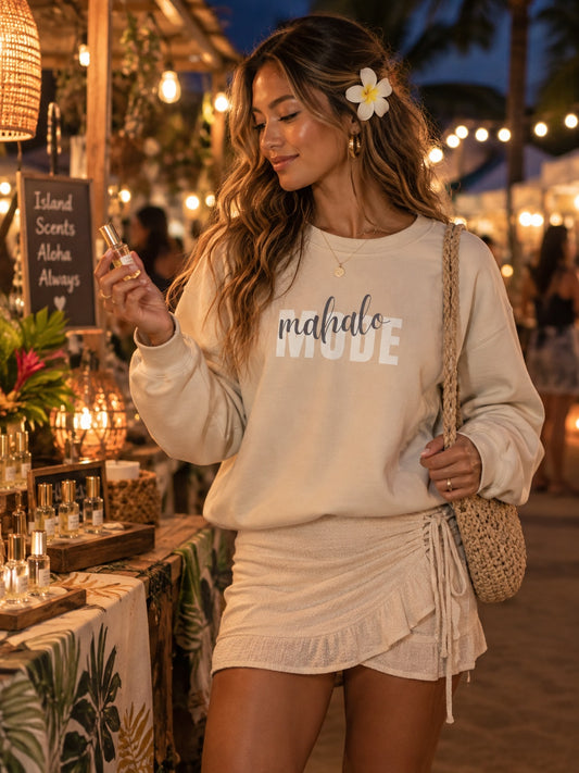 A woman at an outdoor night market wears a cream sweatshirt with 'malaho MODE' printed on the front and a matching cream wrap skirt. She has a white flower in her hair, a woven shoulder bag, and is holding a perfume bottle.