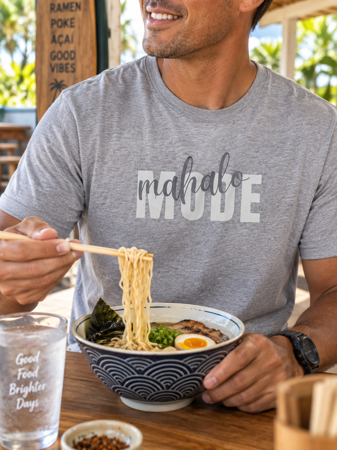 Man wearing a grey Mahalo Mode graphic t-shirt while eating ramen at a casual Hawaiʻi restaurant.