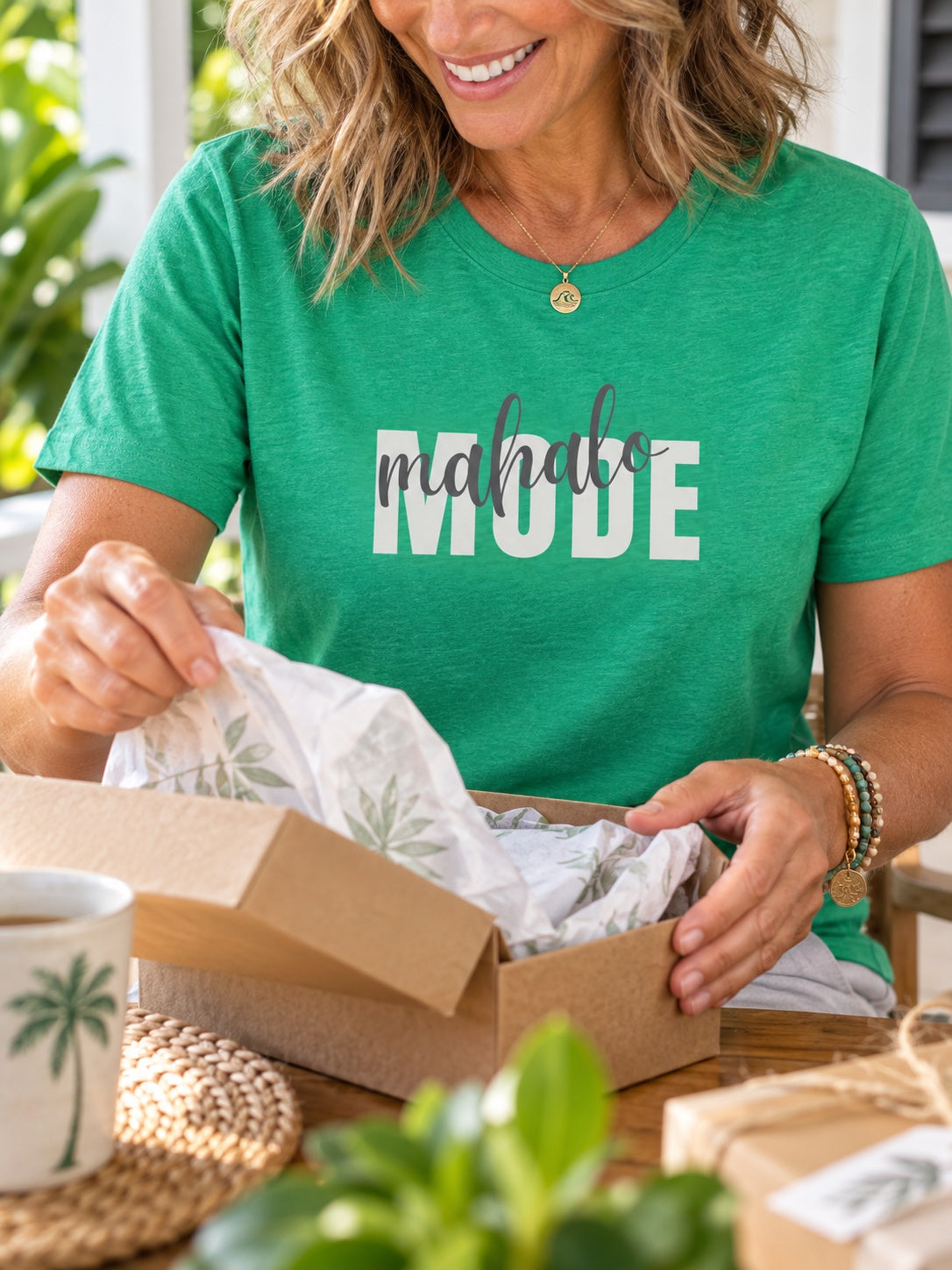 Woman wearing a green Mahalo Mode graphic t-shirt while opening a gift box at home in Hawaiʻi.